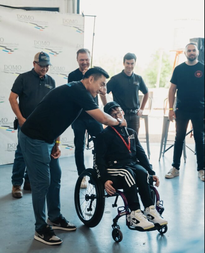 A man handing a microphone to a young person in a wheelchair in front of a DOMO Motorsports Club backdrop, with attendees looking on